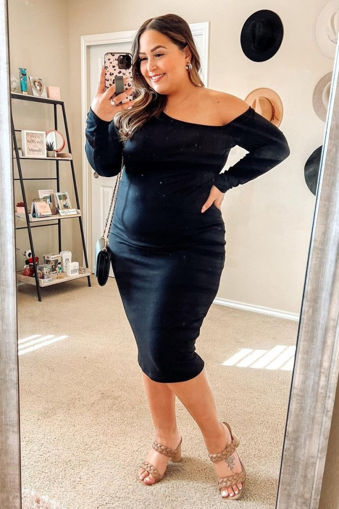 woman wearing a black off-the-shoulder bodycon dress with strappy heels, standing in front of a mirror