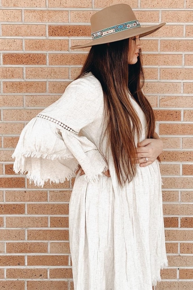 woman wearing a cream maternity dress with fringe sleeves and holding her baby bump, paired with a wide-brimmed hat