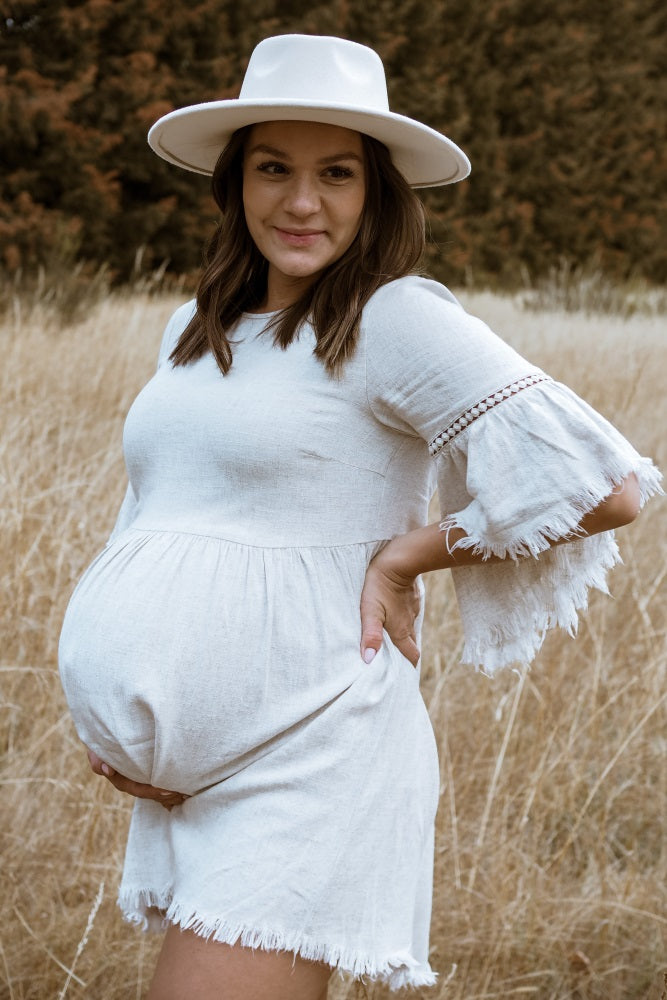 woman wearing a light beige maternity dress with fringe and bell sleeves, showcasing her baby bump in a field