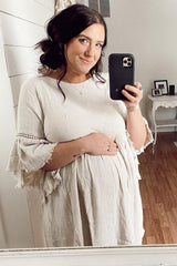 woman wearing a beige maternity dress with fringe details, holding her baby bump in a mirror selfie