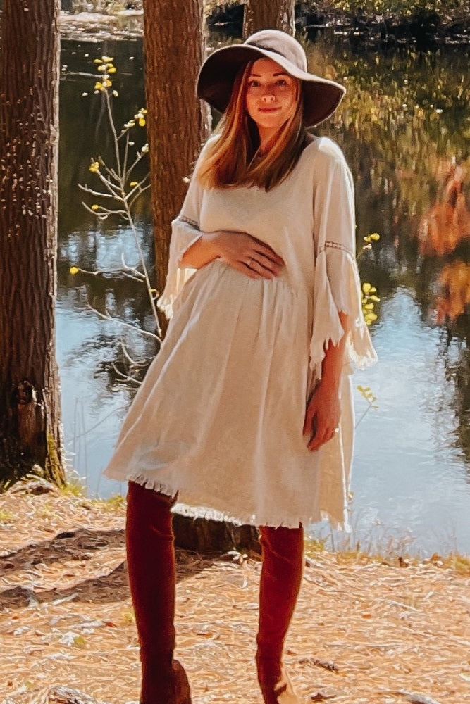 woman wearing a cream maternity dress with flowy sleeves and a baby bump, accessorized with a wide-brimmed hat and tall boots