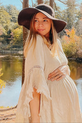 woman wearing a beige maternity dress with fringed sleeves and a wide-brim hat, gently holding her baby bump outdoors