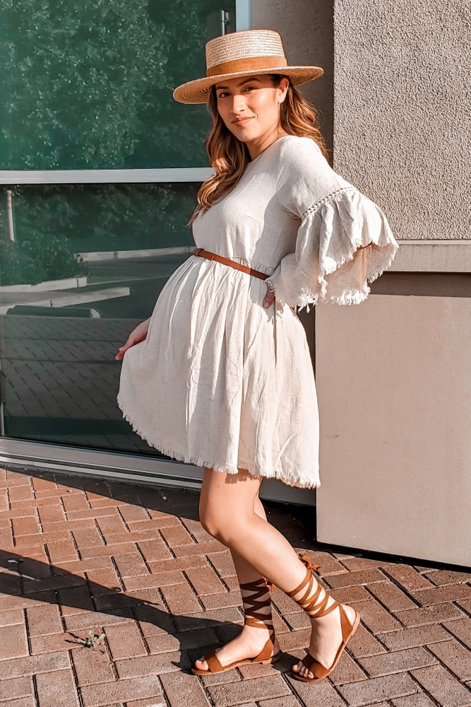 woman wearing a beige maternity dress with bell sleeves and a brown belt, showcasing her baby bump, paired with strappy sandals and a hat