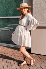 woman wearing a beige maternity dress with bell sleeves and a brown belt, showcasing her baby bump, paired with strappy sandals and a hat