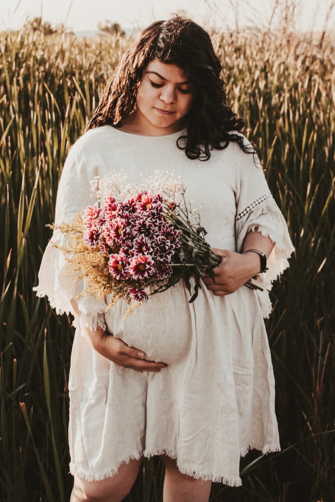 woman wearing a beige maternity dress with lace details, holding a bouquet and cradling her baby bump in a field