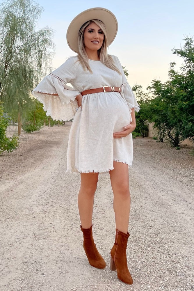 woman wearing a cream maternity dress with bell sleeves, belted at the waist, and a baby bump visible, paired with brown high-heeled boots and a wide-brimmed hat