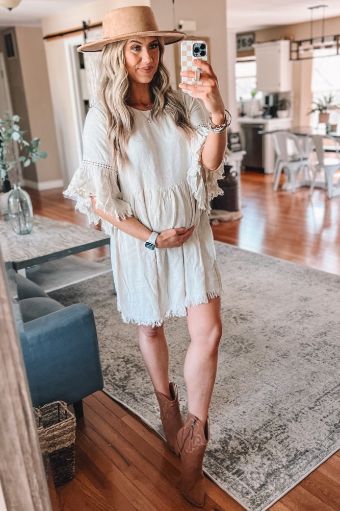 woman wearing a light beige maternity dress with ruffle sleeves and holding her baby bump, paired with a wide-brim hat and boots