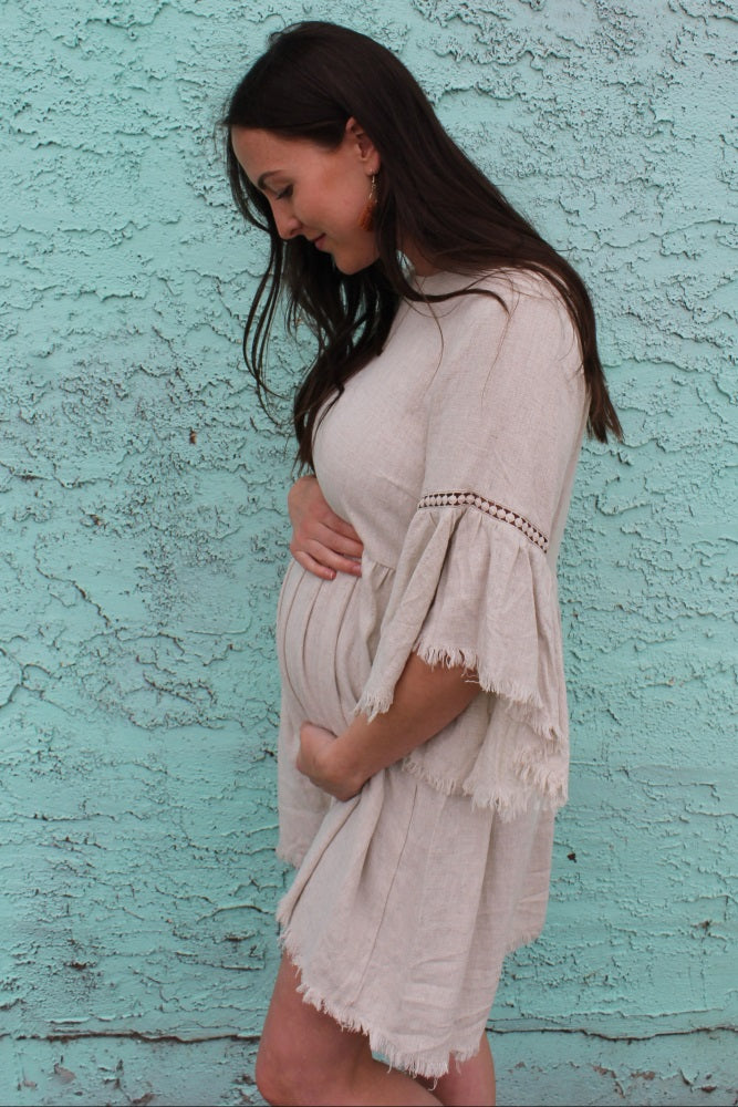 woman wearing a beige maternity dress with bell sleeves and holding her baby bump against a turquoise backdrop