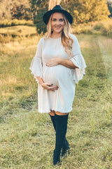 woman wearing a light beige maternity dress with bell sleeves and holding her baby bump in a grassy field