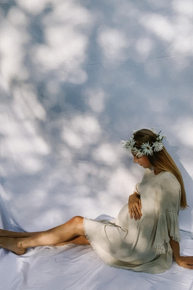 woman wearing a cream colored maternity dress with tassel details and a flower crown, holding her baby bump while seated