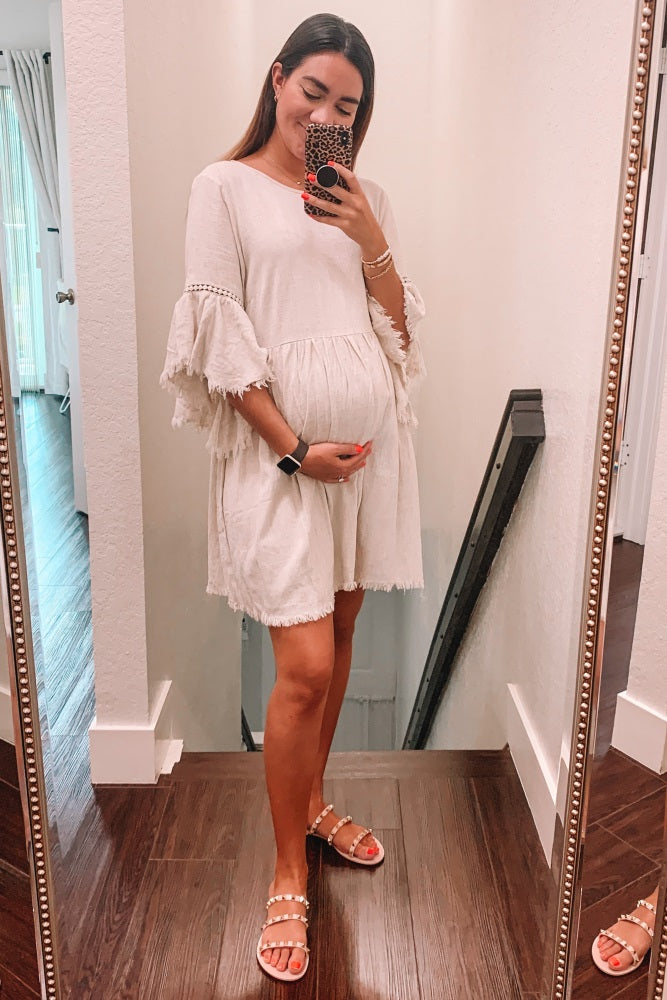 woman wearing a cream maternity dress with ruffled sleeves, showing her baby bump while standing in front of a mirror
