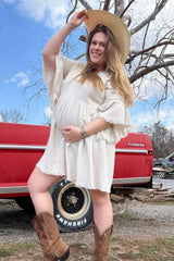 woman wearing a cream maternity dress with fringe details and a large hat, holding her baby bump outdoors