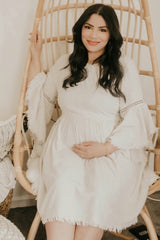 woman wearing a cream maternity dress with bell sleeves, sitting in a hanging chair and holding her baby bump