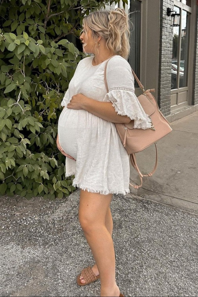 woman wearing a white maternity dress with fringed sleeves and holding a pink bag, showing her baby bump