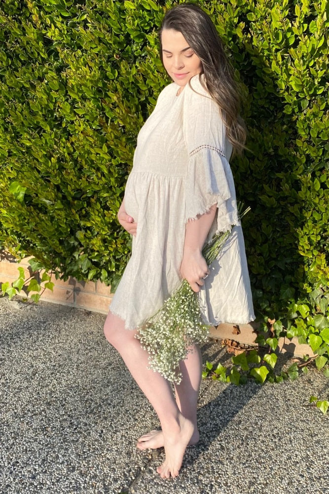 woman wearing a light beige maternity dress with short sleeves and a flowing silhouette, holding a bouquet of flowers and showing her baby bump