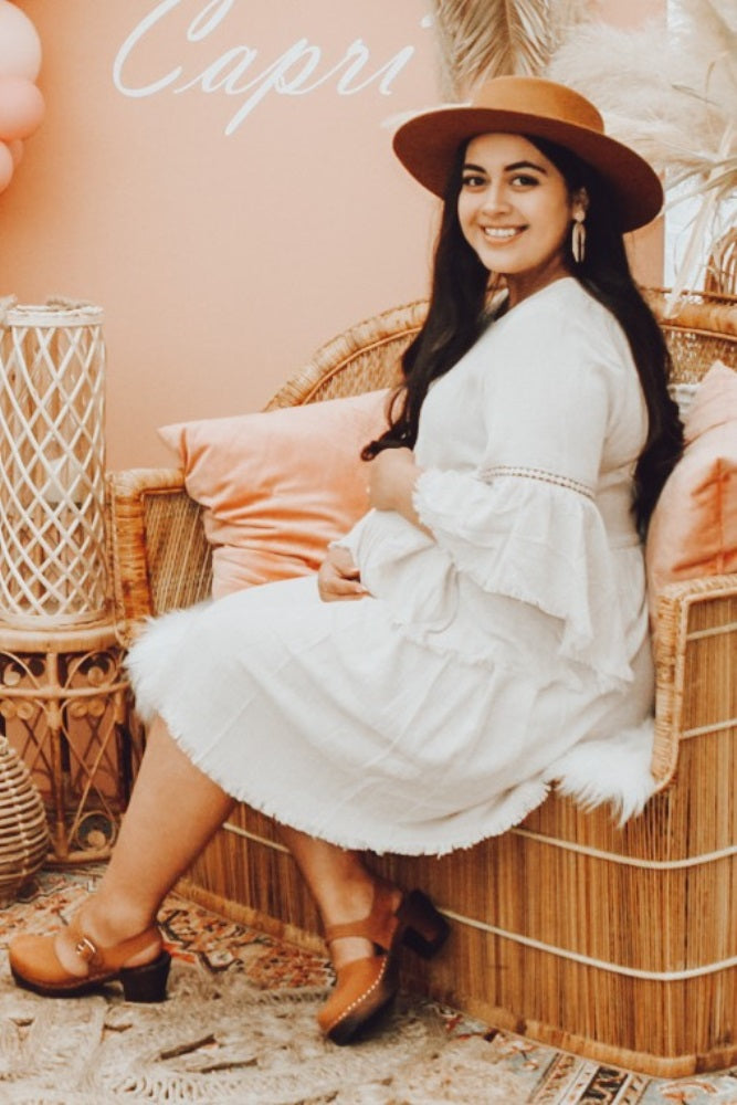 Smiling pregnant woman in white dress and brown hat sitting on a wicker chair at a baby shower