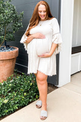 woman wearing a light beige short sleeve maternity dress and holding her baby bump outside.