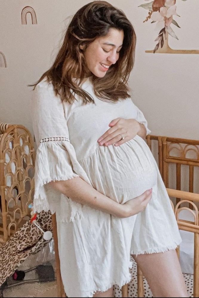 woman wearing a light beige maternity dress with ruffled sleeves, holding her baby bump standing in a nursery
