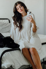 woman wearing a white maternity dress with frayed edges and bell sleeves, sitting on a bed, holding a phone, dog beside her