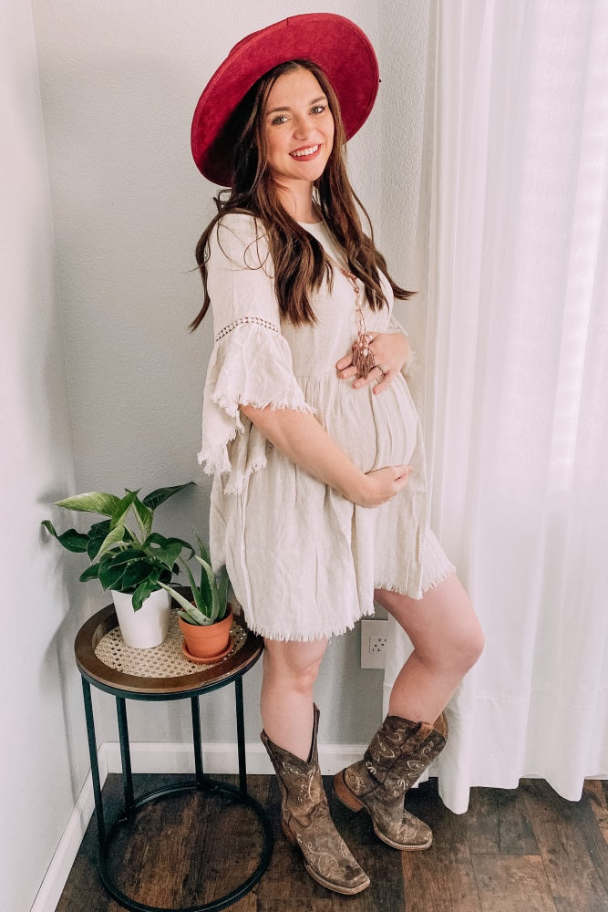 woman wearing a light beige maternity dress and cowboy boots, holding her baby bump while smiling