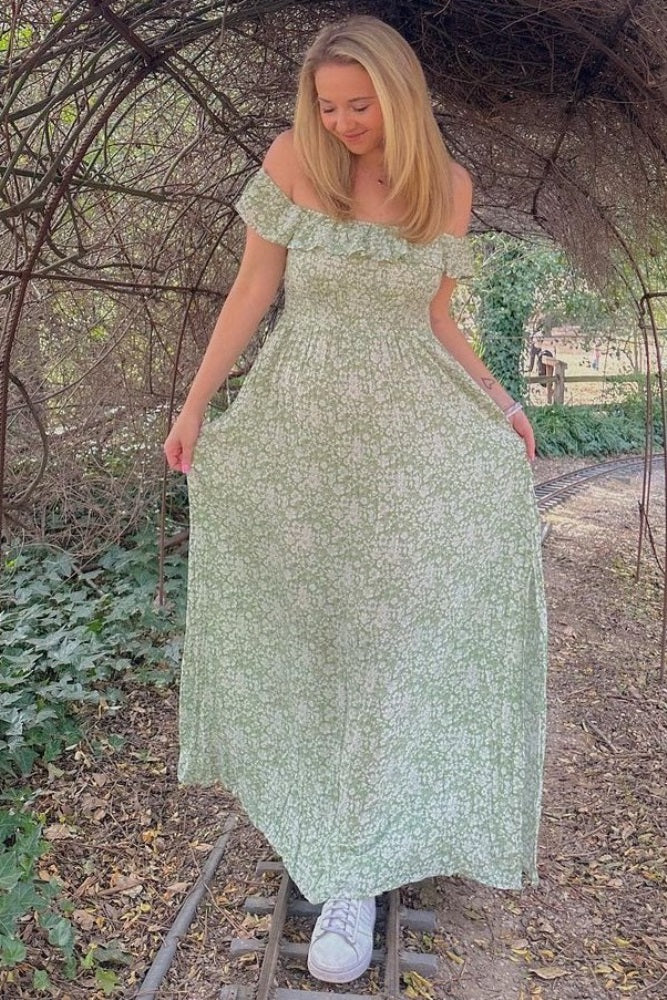 woman wearing a green off-the-shoulder floral maxi dress and white sneakers, posing outdoors among greenery