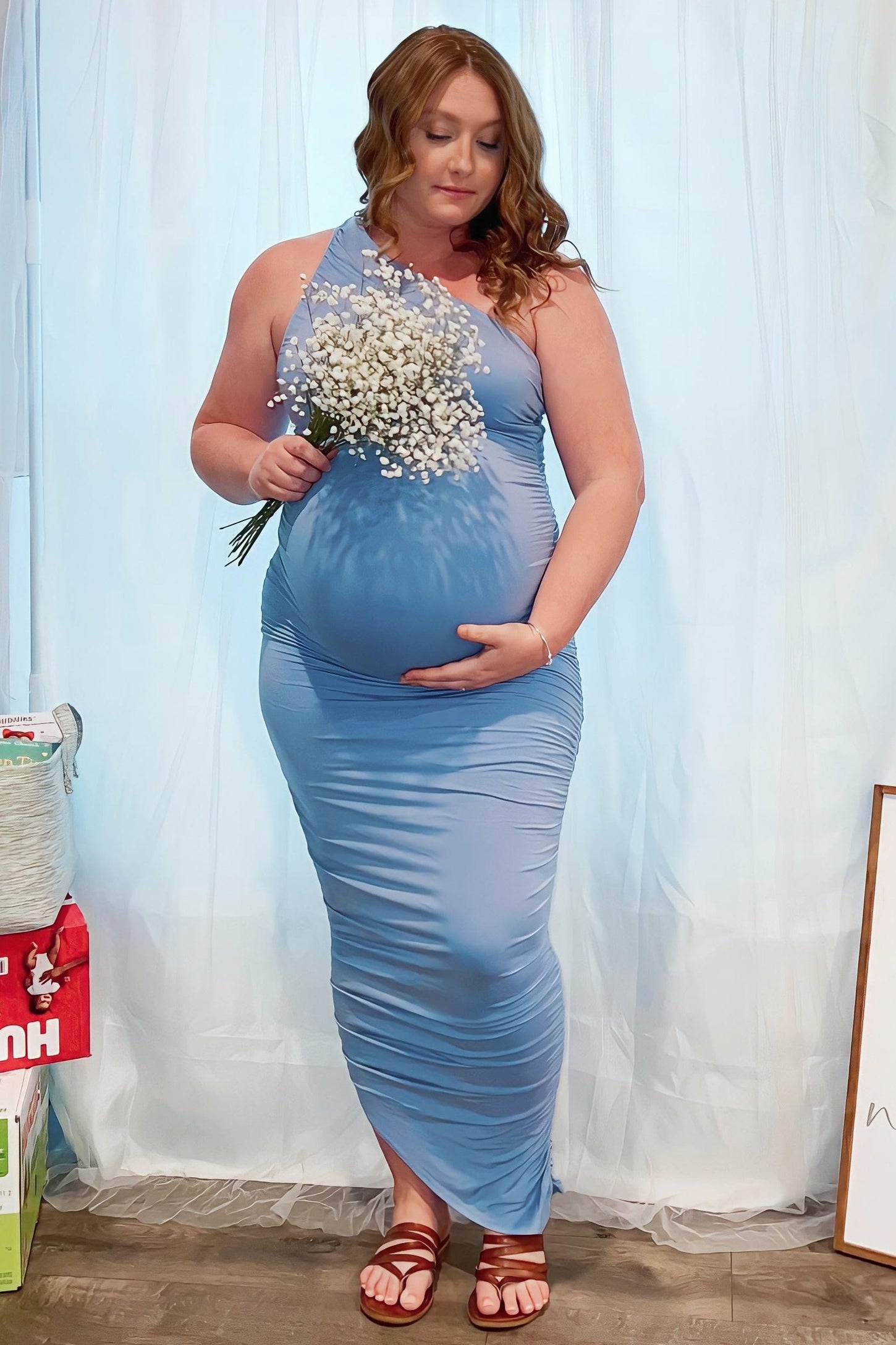 woman wearing a light blue one-shoulder maternity dress holding flowers and showing her baby bump