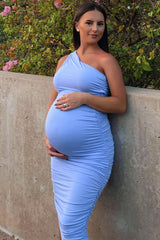 woman wearing a light blue one-shoulder maternity dress, gently cradling her baby bump while leaning against a wall