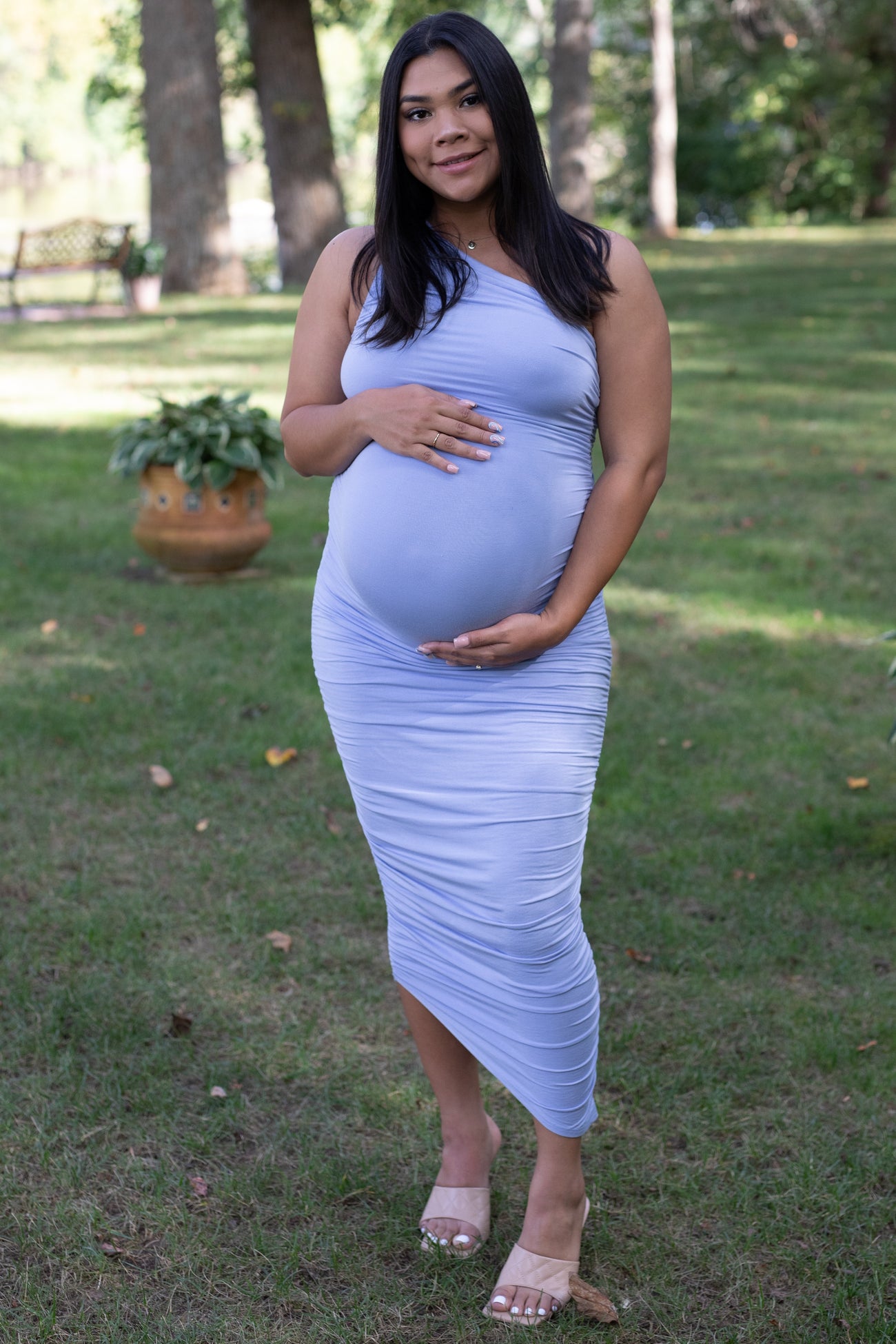 woman wearing a light lavender maternity dress, cradling her baby bump in a grassy outdoor setting