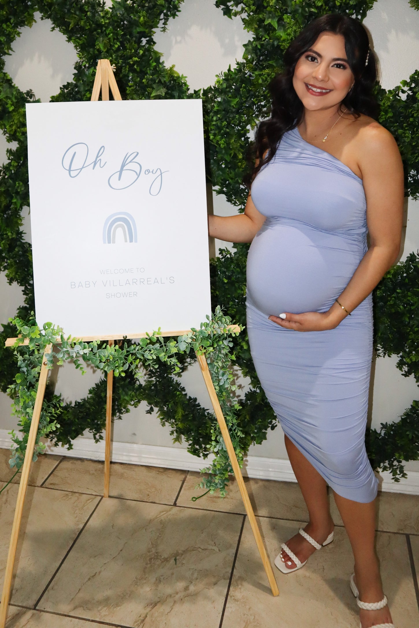 woman wearing a light blue fitted maternity dress with a ruffled shoulder, holding her baby bump