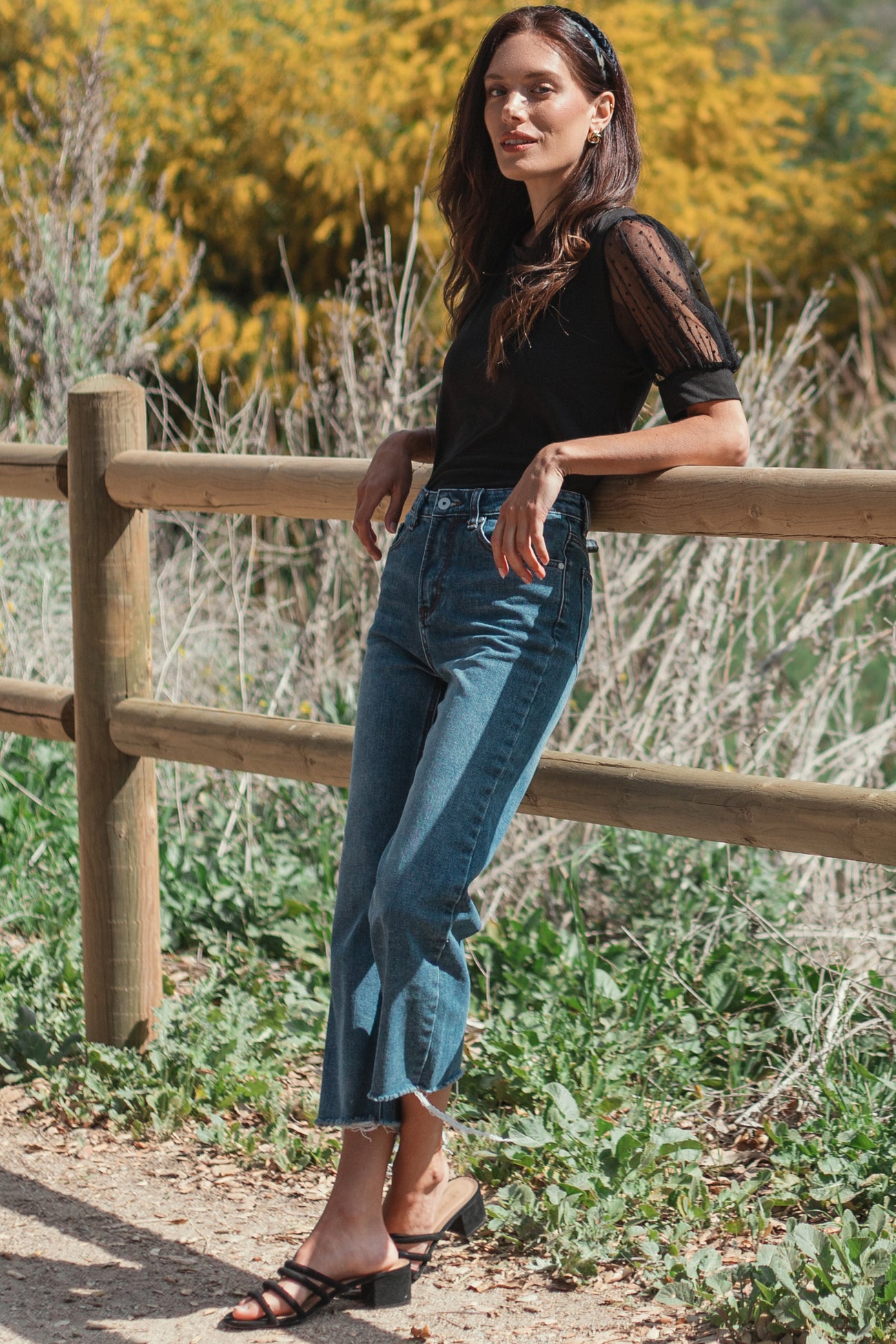 woman wearing a black sheer sleeve top and high-waisted blue jeans, standing by a wooden fence outdoors