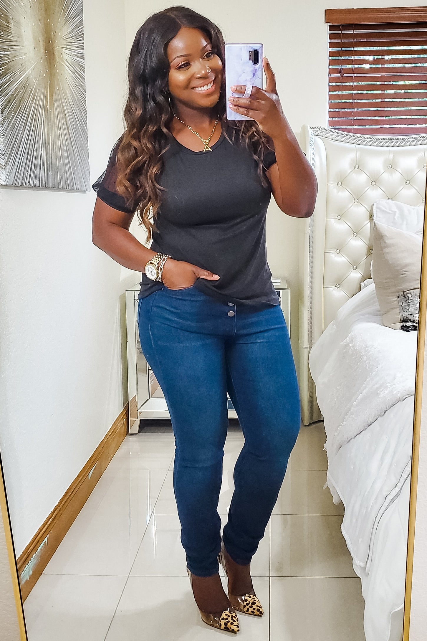 woman wearing a black short sleeve top and blue skinny jeans, smiling while posing in front of a mirror