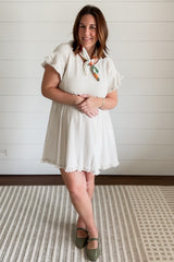 woman wearing a light beige short sleeve dress with ruffled sleeves and a colorful scarf, standing on a patterned rug