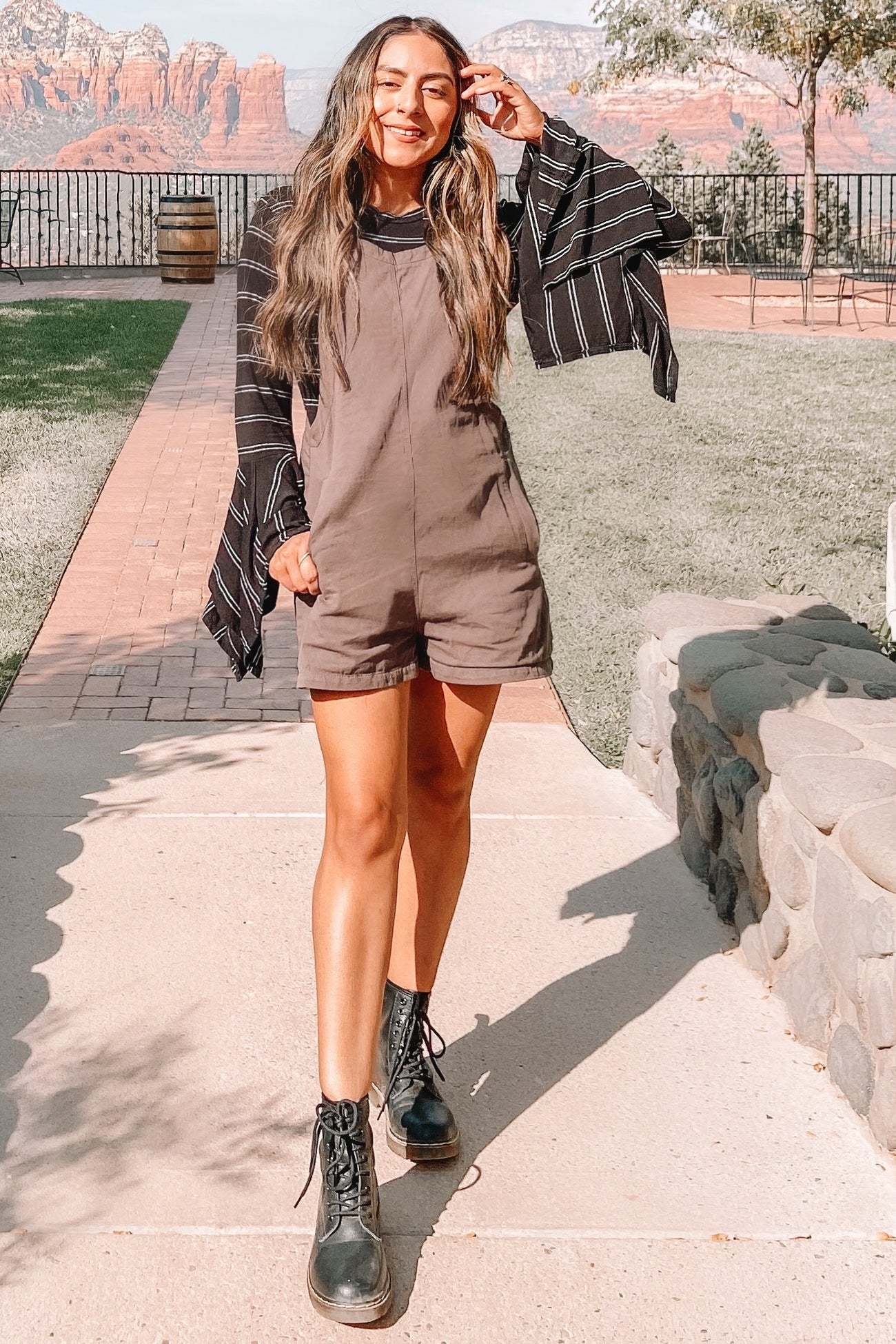woman in brown short overalls and black combat boots standing on a path with red rock mountains in the background