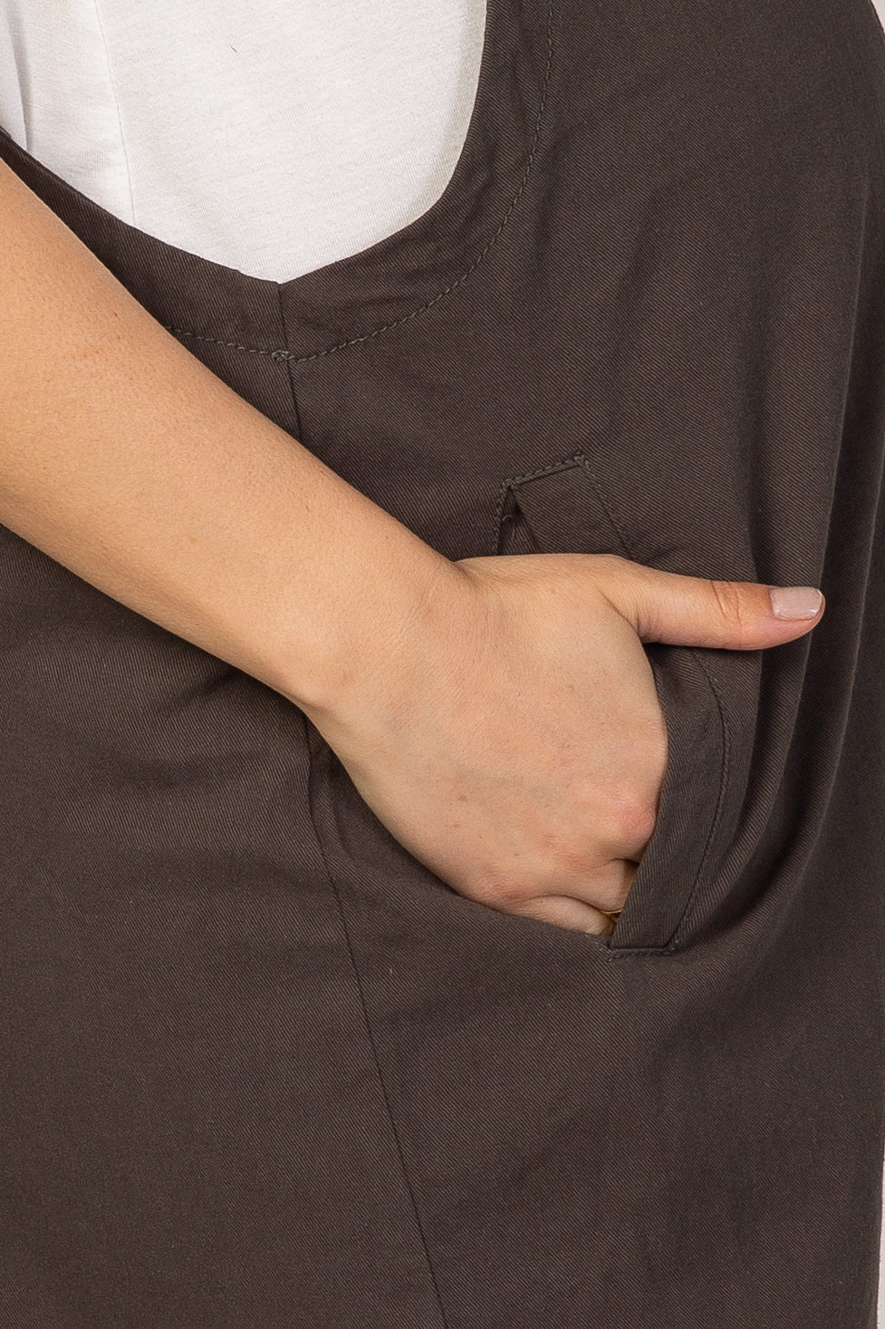 Close up of a hand in the side pocket of dark brown cotton overalls worn over a white t-shirt