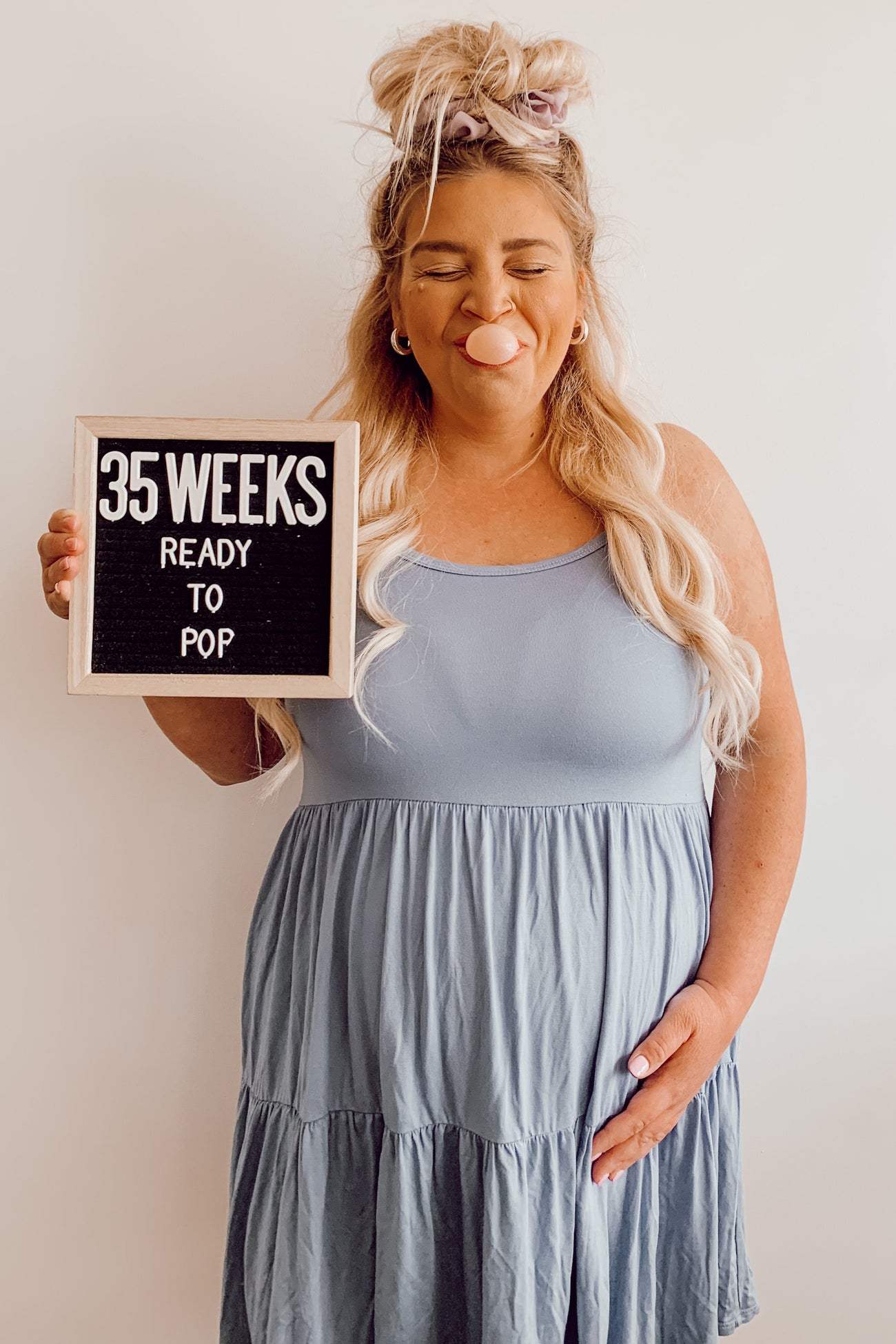 woman wearing a light blue sleeveless maternity dress holding a sign that says "35 weeks ready to pop" with a visible baby bump