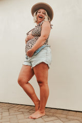 woman wearing a leopard print maternity swimsuit and denim shorts, holding her baby bump and smiling