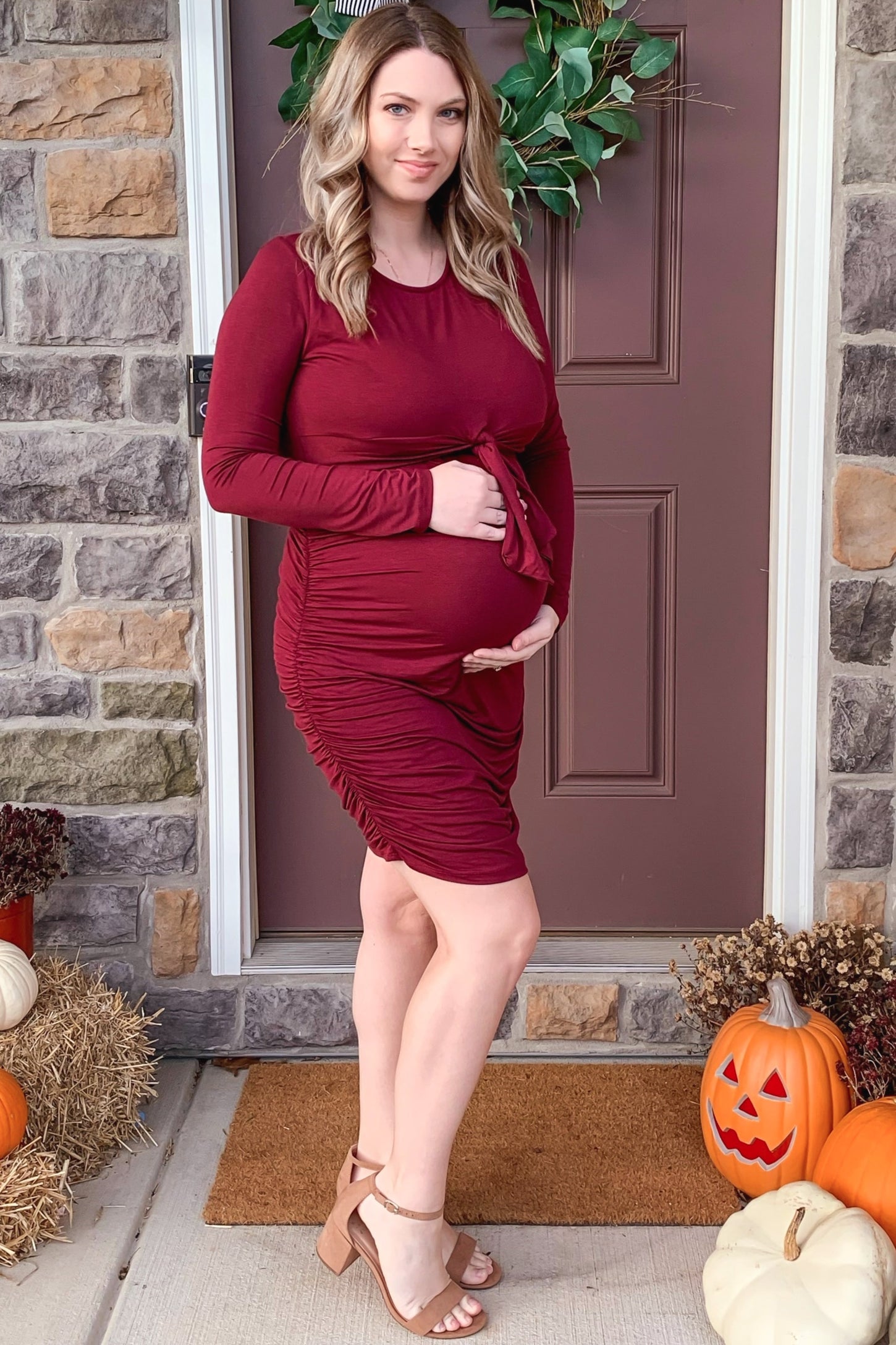 woman wearing a burgundy maternity dress with ruching, holding her baby bump while standing by a front door