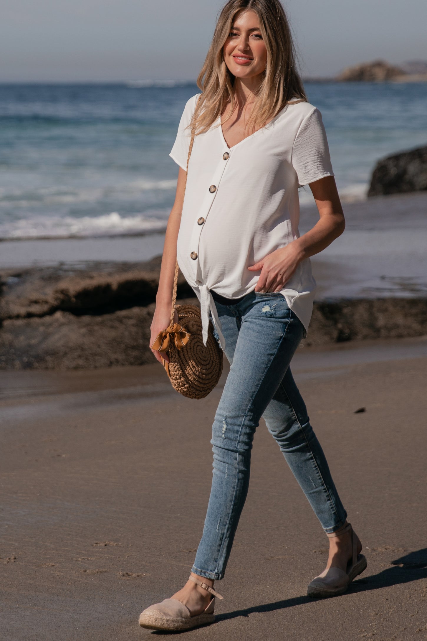 woman wearing a white short sleeve maternity top with buttons and tie front, paired with distressed blue skinny jeans and holding a straw bag
