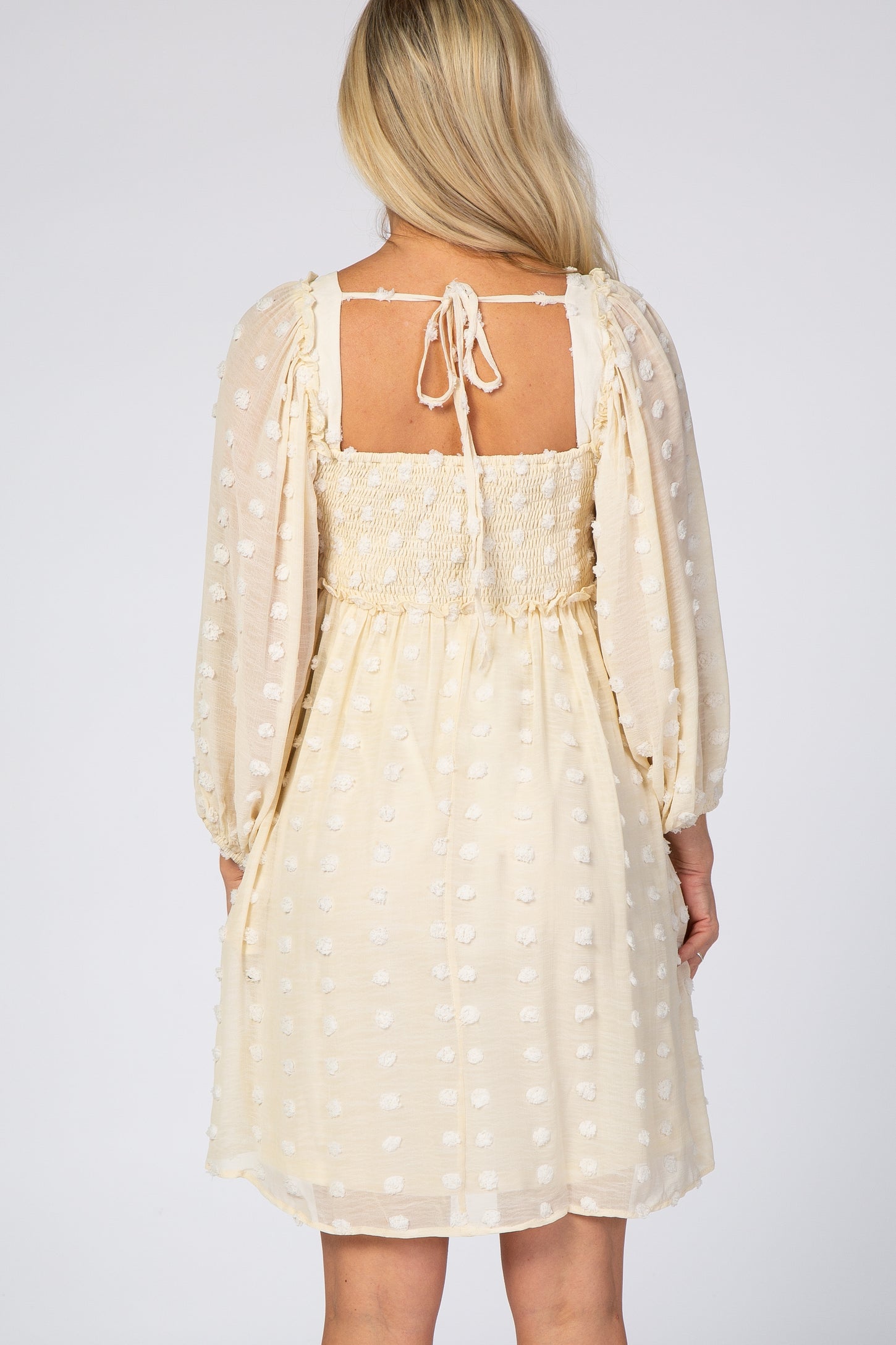 Back view of a woman wearing a cream swiss dot maternity dress with long puff sleeves and a smocked back.
