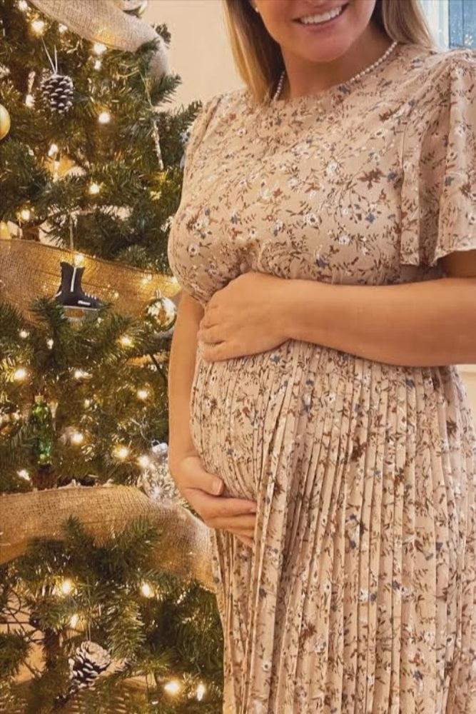 woman wearing a floral pleated maternity dress and holding her baby bump in front of a decorated Christmas tree