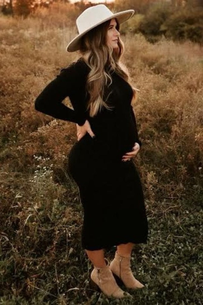 pregnant woman in black midi dress and white wide brim hat standing in a field at sunset
