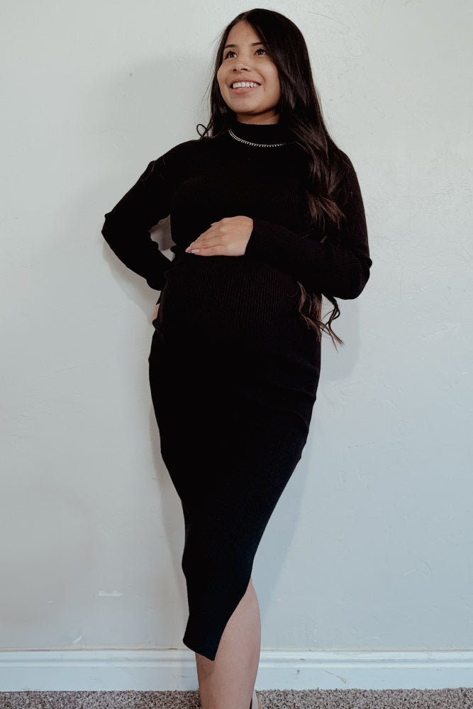 Smiling pregnant woman in a black long-sleeve maternity midi dress holding her baby bump