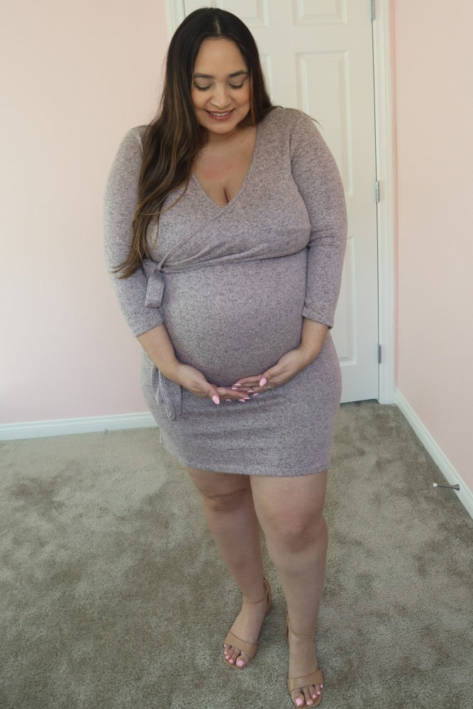 Pregnant woman in a mauve wrap maternity dress holding her baby bump in a room with pink walls