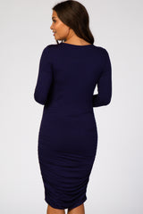 Back view of a woman wearing a long sleeve navy blue ruched maternity dress against a white background