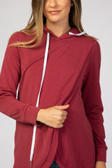 woman wearing a burgundy maternity hoodie with a white drawstring and long sleeves, gently holding her baby bump