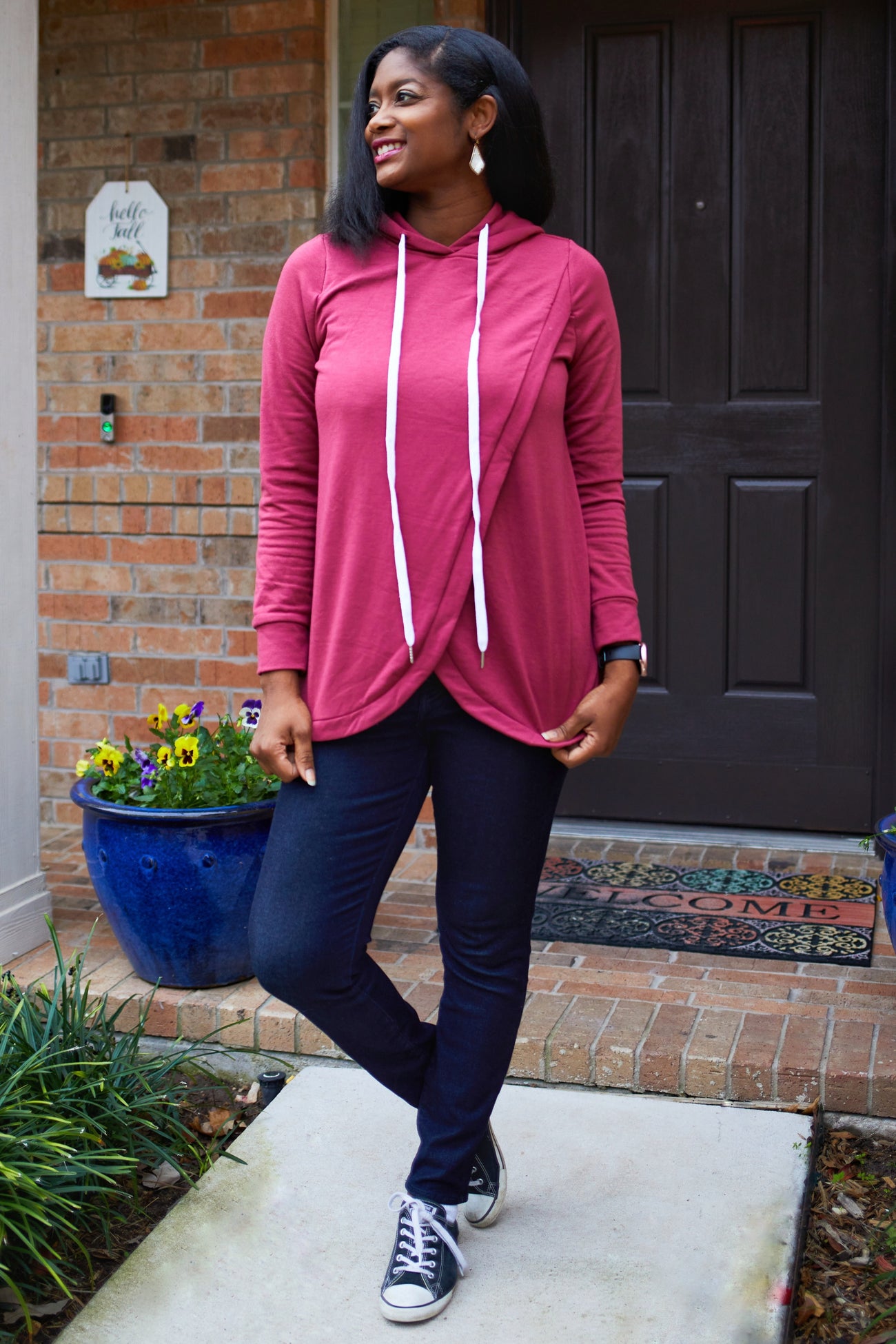 woman wearing a maroon hooded long sleeve top with white drawstrings and dark jeans, standing outside a house