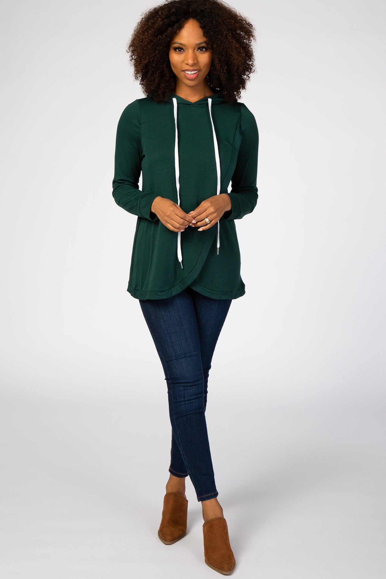woman wearing a green long sleeve hooded top with drawstrings and dark skinny jeans, standing in brown ankle boots