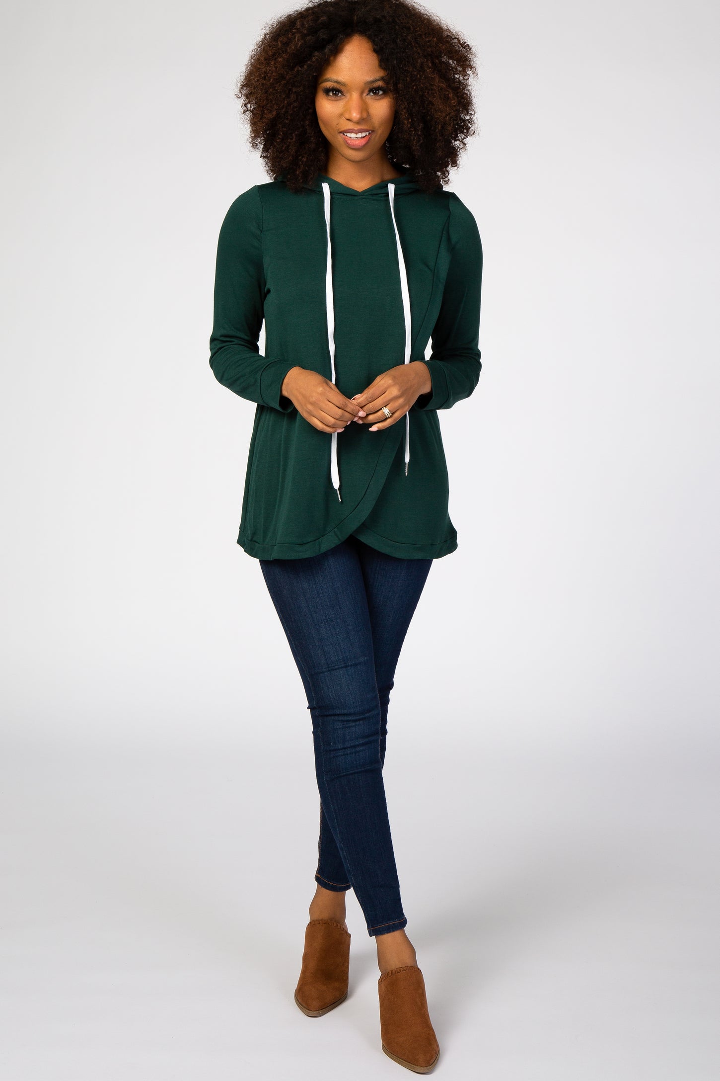 woman wearing a green long sleeve hooded top with drawstrings and dark skinny jeans, standing in brown ankle boots