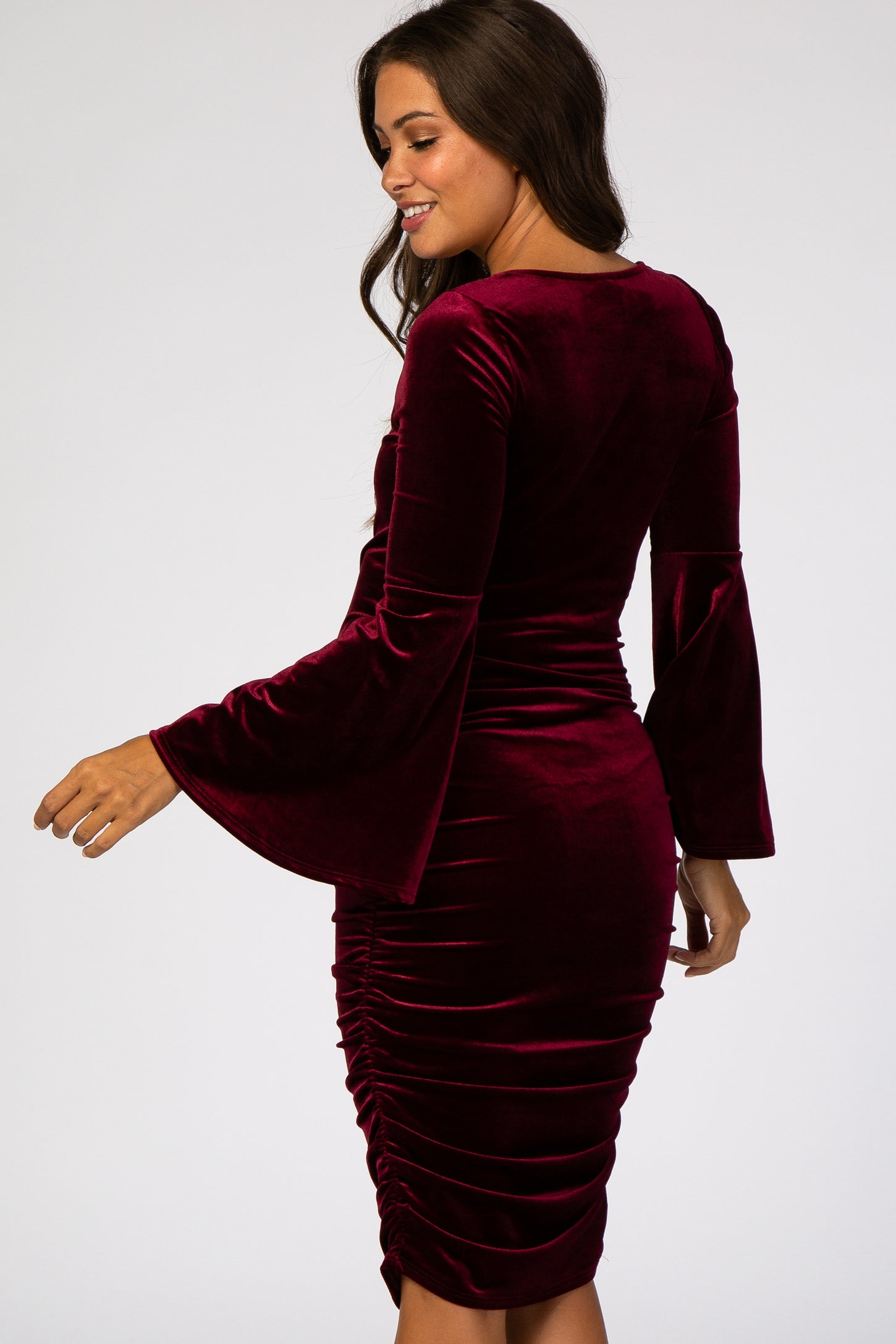 woman wearing a burgundy velvet maternity dress with bell sleeves and ruching detail, showing off her baby bump