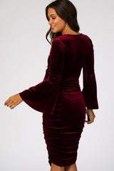 woman wearing a burgundy velvet maternity dress with bell sleeves and ruching detail, showing off her baby bump